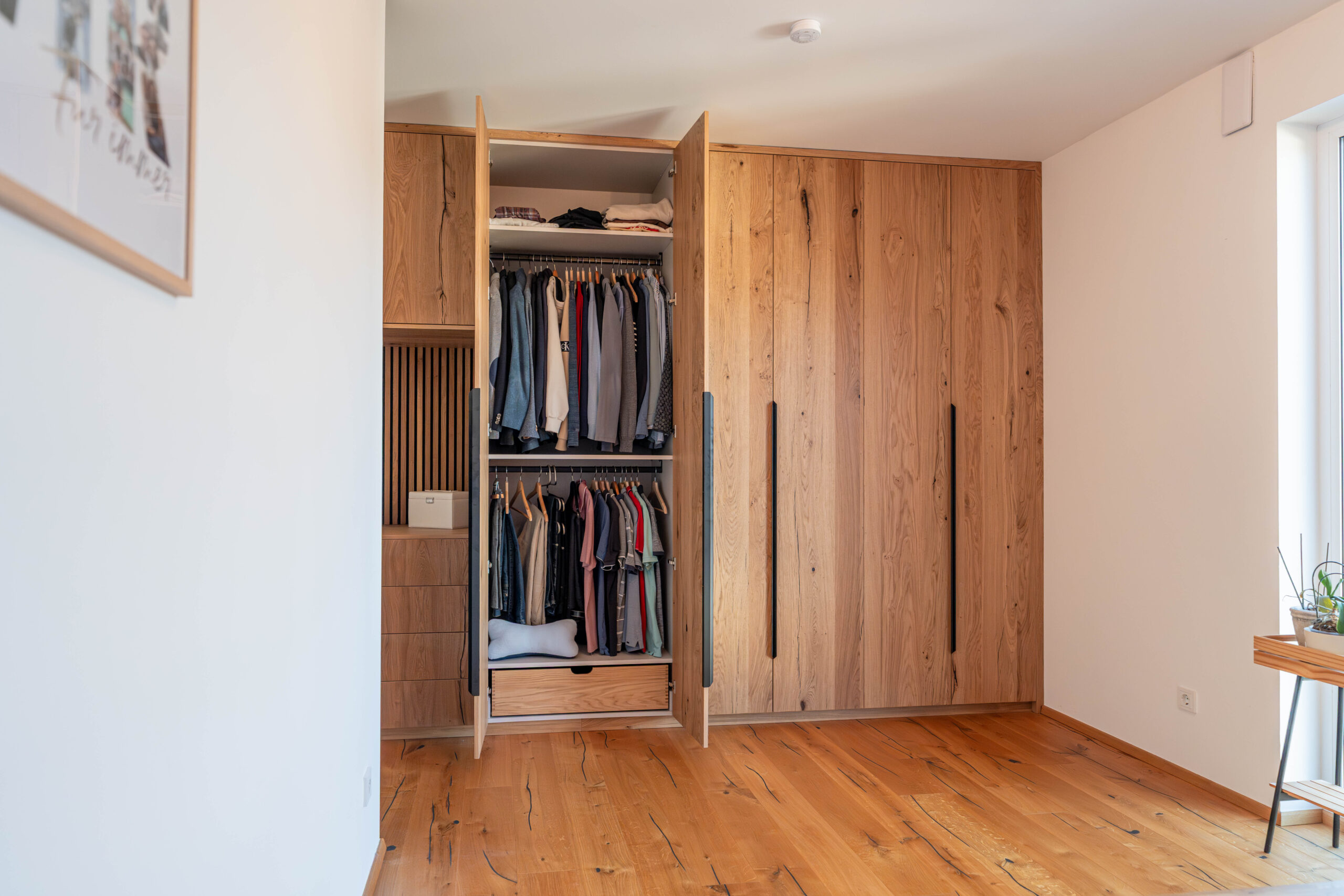 Open, custom-made bedroom closet made of rustic oak with two clothing rods, drawers, and black handle strips.