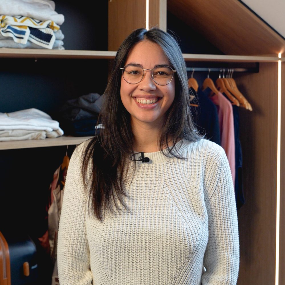Kundin Jennifer W. vor ihrem geöffneten maßgefertigten Dachschrägenschrank mit Kleiderstange, Regalfächern und LED-Beleuchtung.